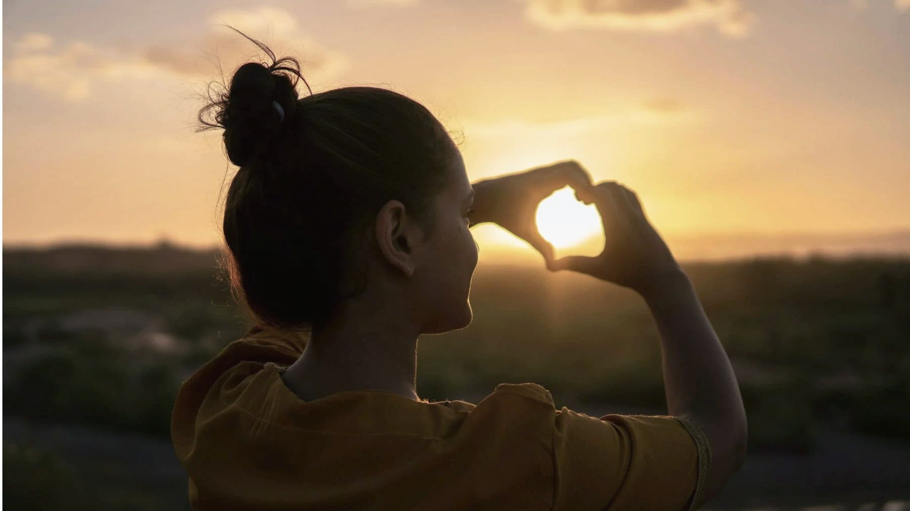 A woman silhouetted against the setting sun, hands raised.